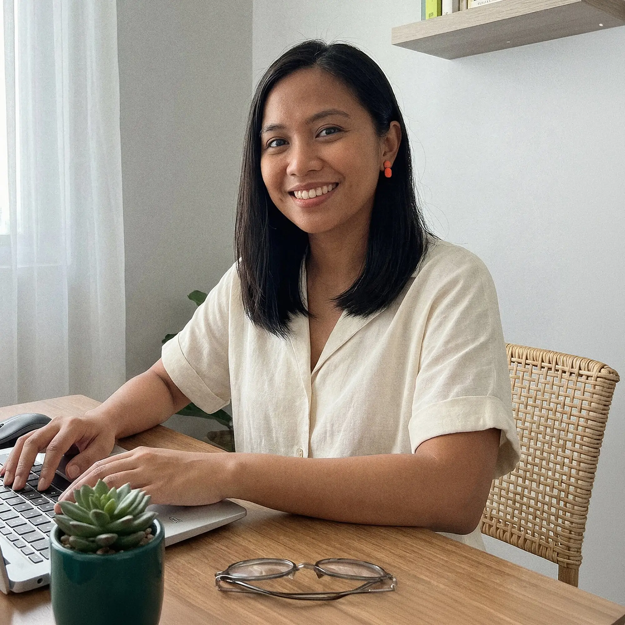 Smiling woman sitting at a wooden desk typing on a laptop with glasses and a small potted succulent nearby.