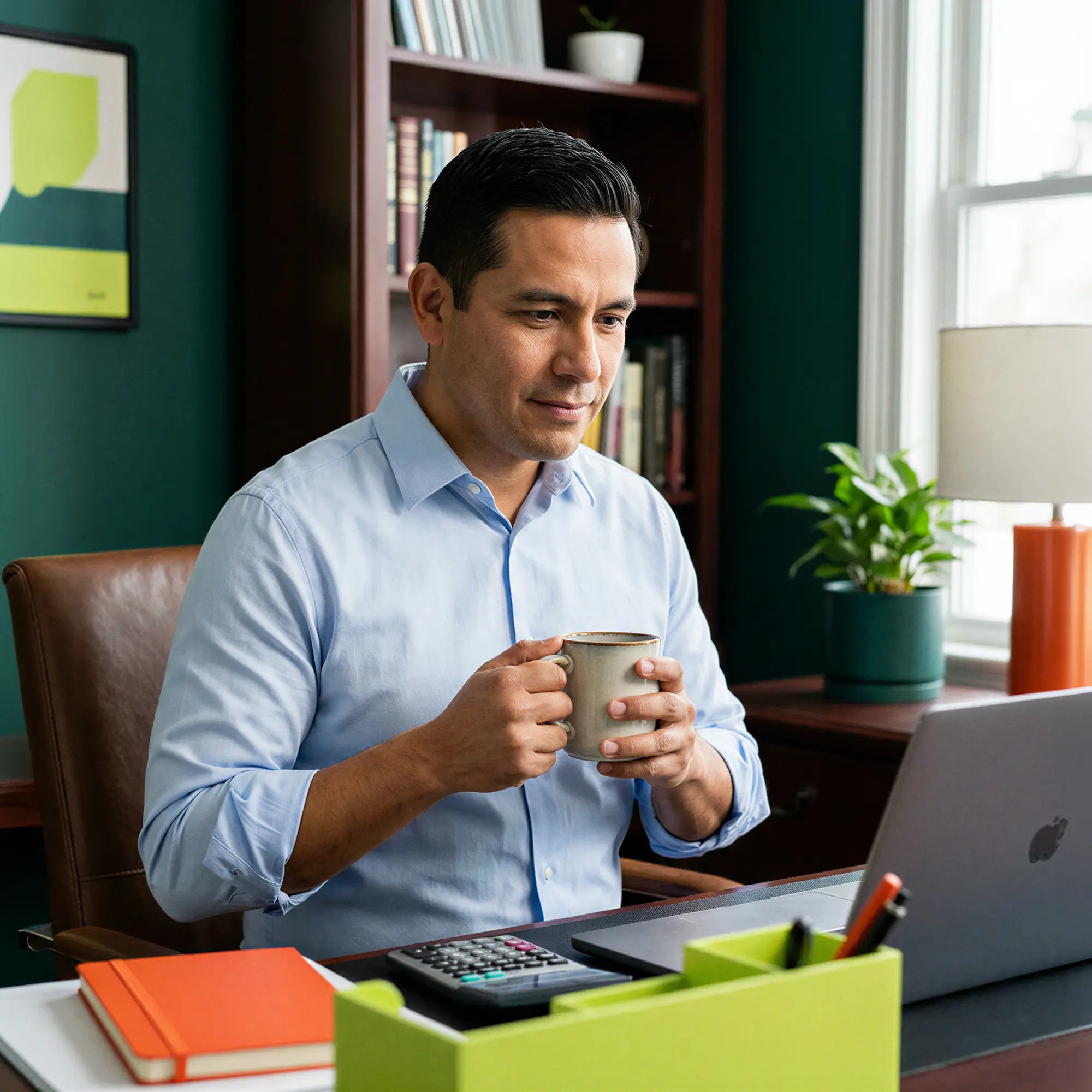 Man in a blue shirt holding a mug while looking at a laptop at his desk.