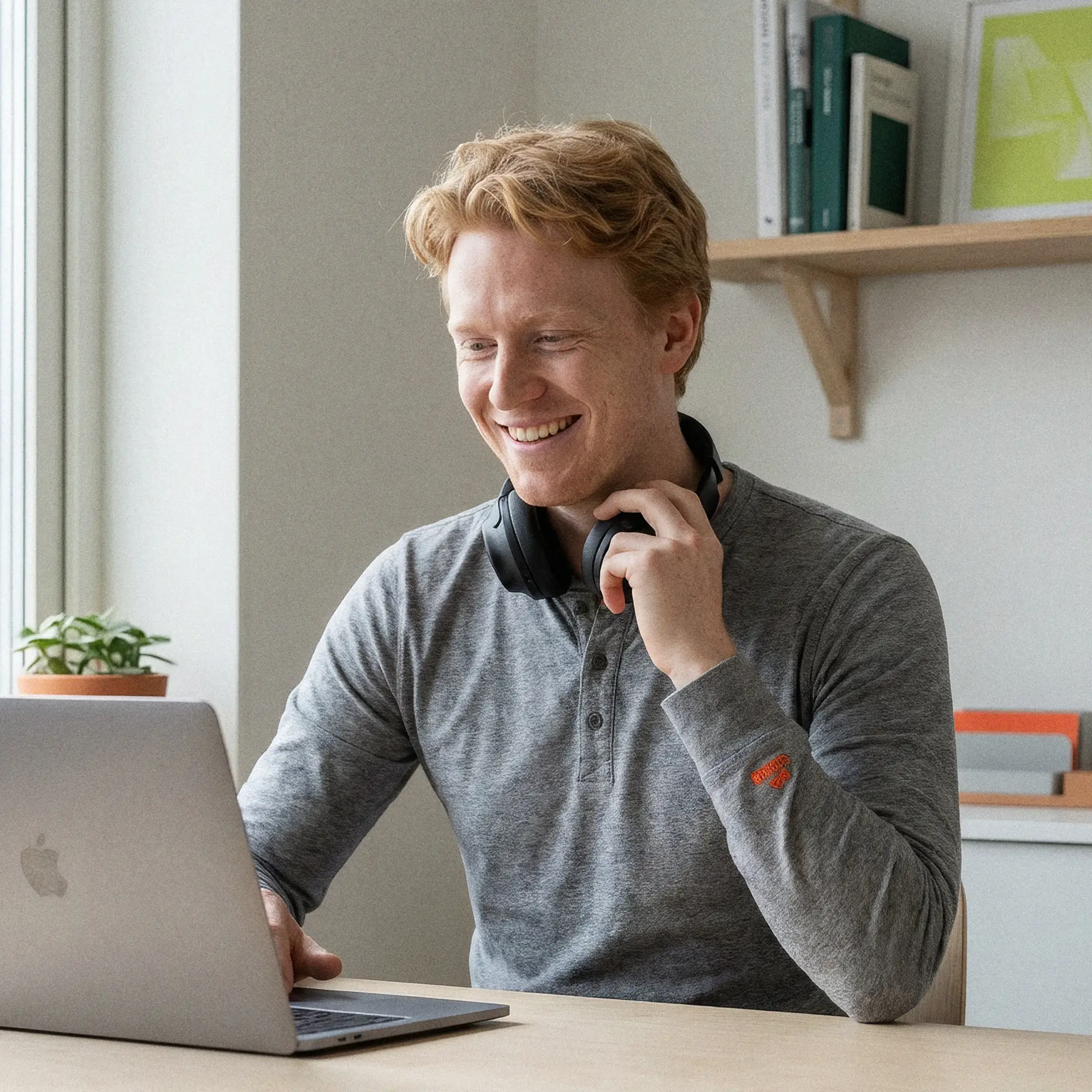 Smiling man with red hair wearing a gray shirt and black headphones around his neck using a laptop at a desk.