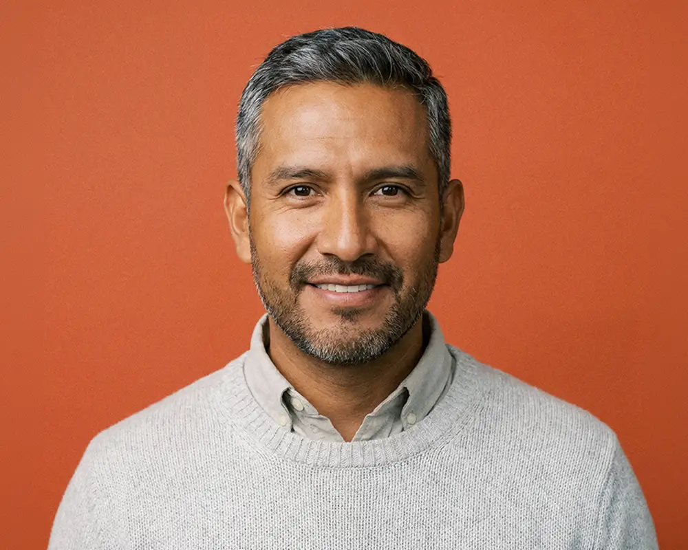 Smiling middle-aged man with salt-and-pepper hair and beard wearing a light grey sweater in front of an orange background.