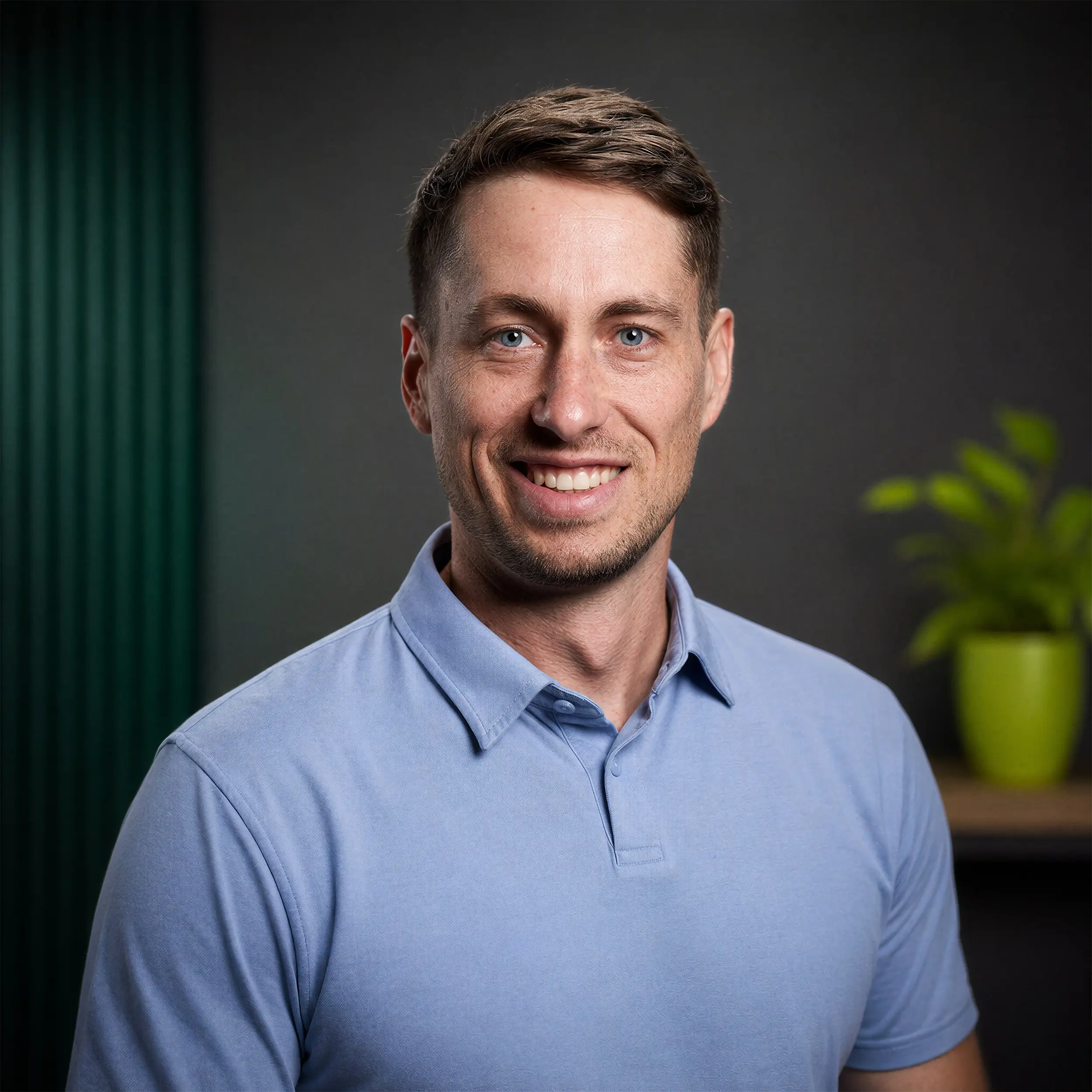 Smiling man with short brown hair wearing a light blue polo shirt against a dark background with a green potted plant.
