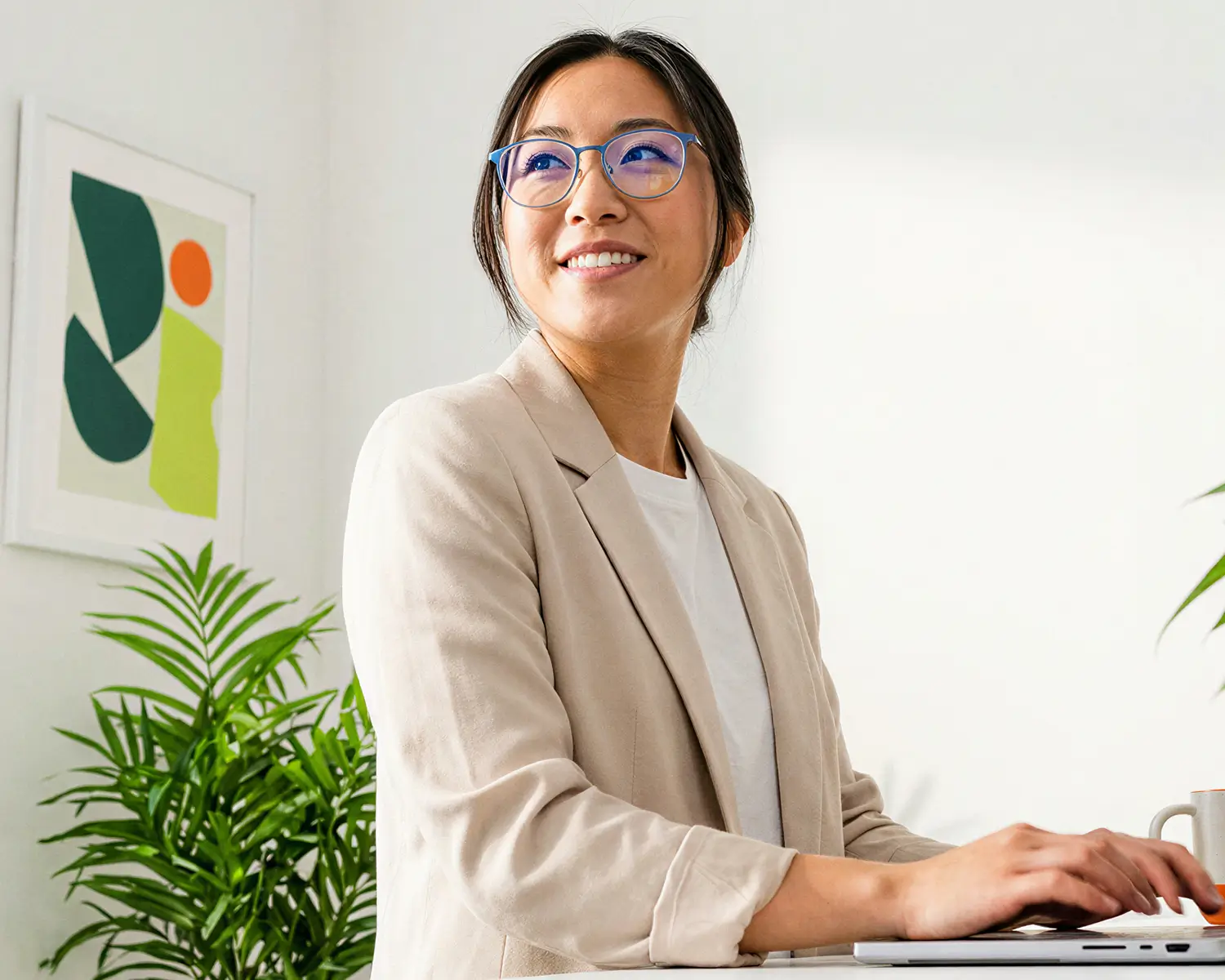 Smiling woman wearing glasses and a beige blazer working on a laptop with green plants and abstract art in the background.