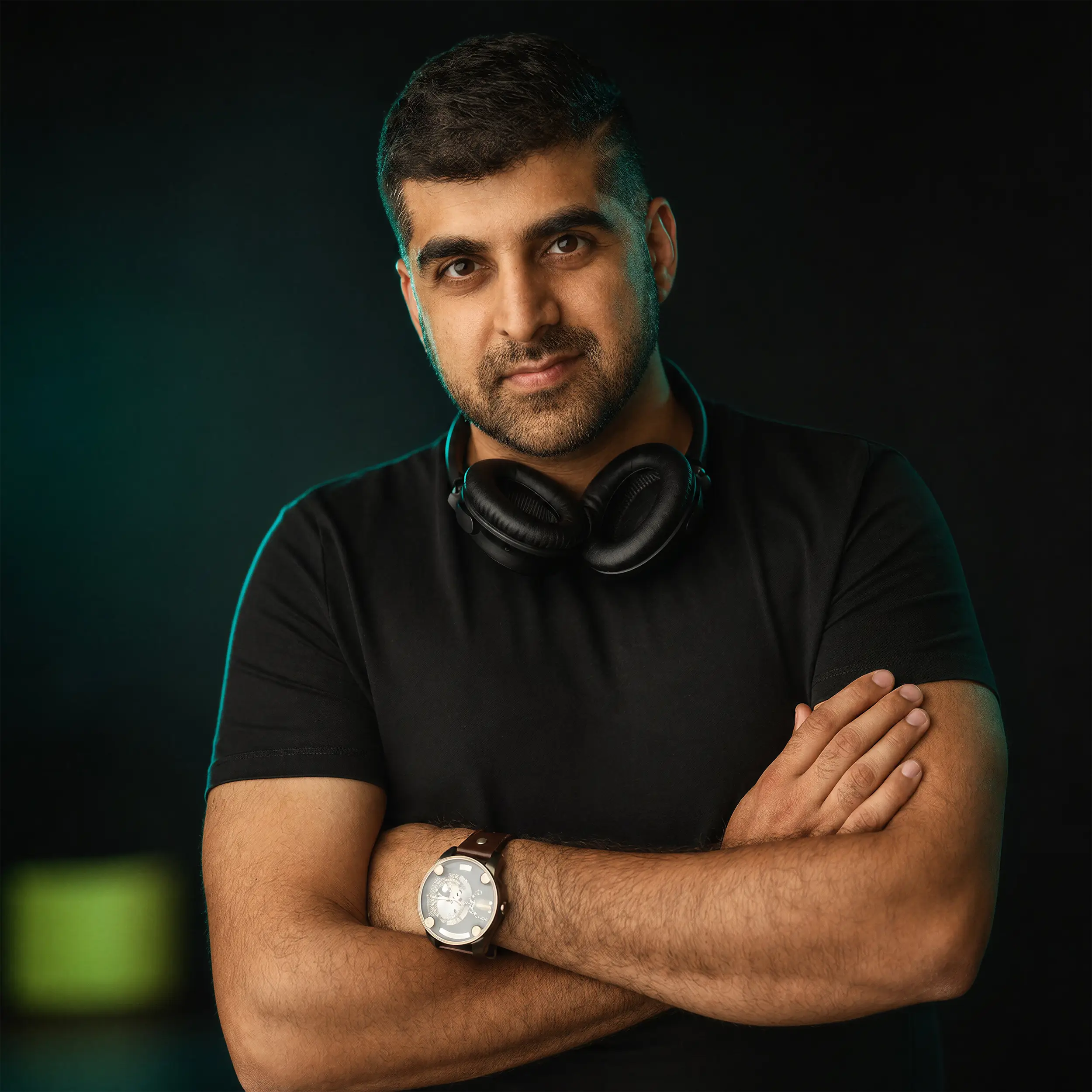 Man with short dark hair wearing a black t-shirt, headphones around his neck, and a watch, posing with arms crossed against a dark background.