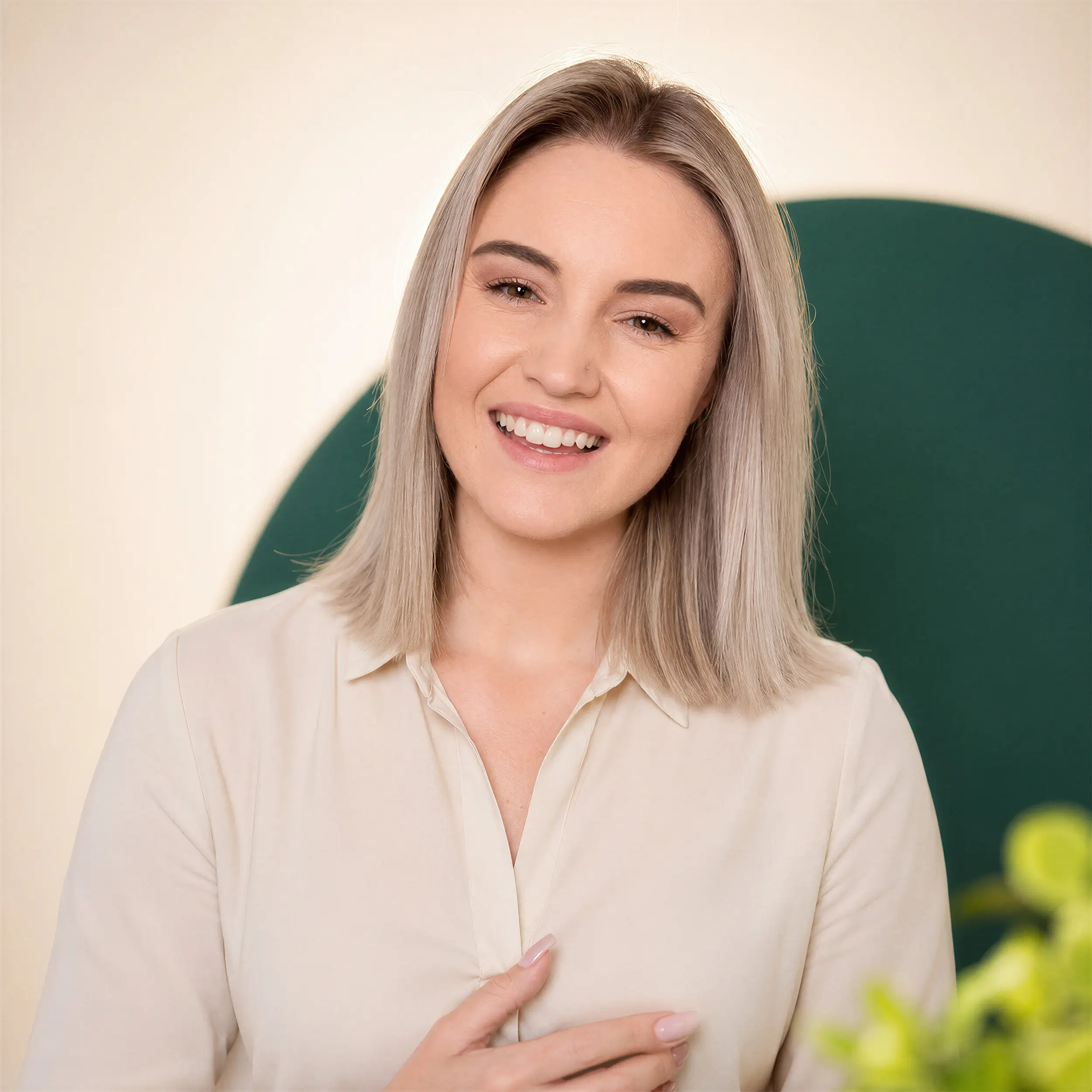 Smiling woman with straight blond hair wearing a light beige blouse against a neutral background with green and yellow blurred plants.