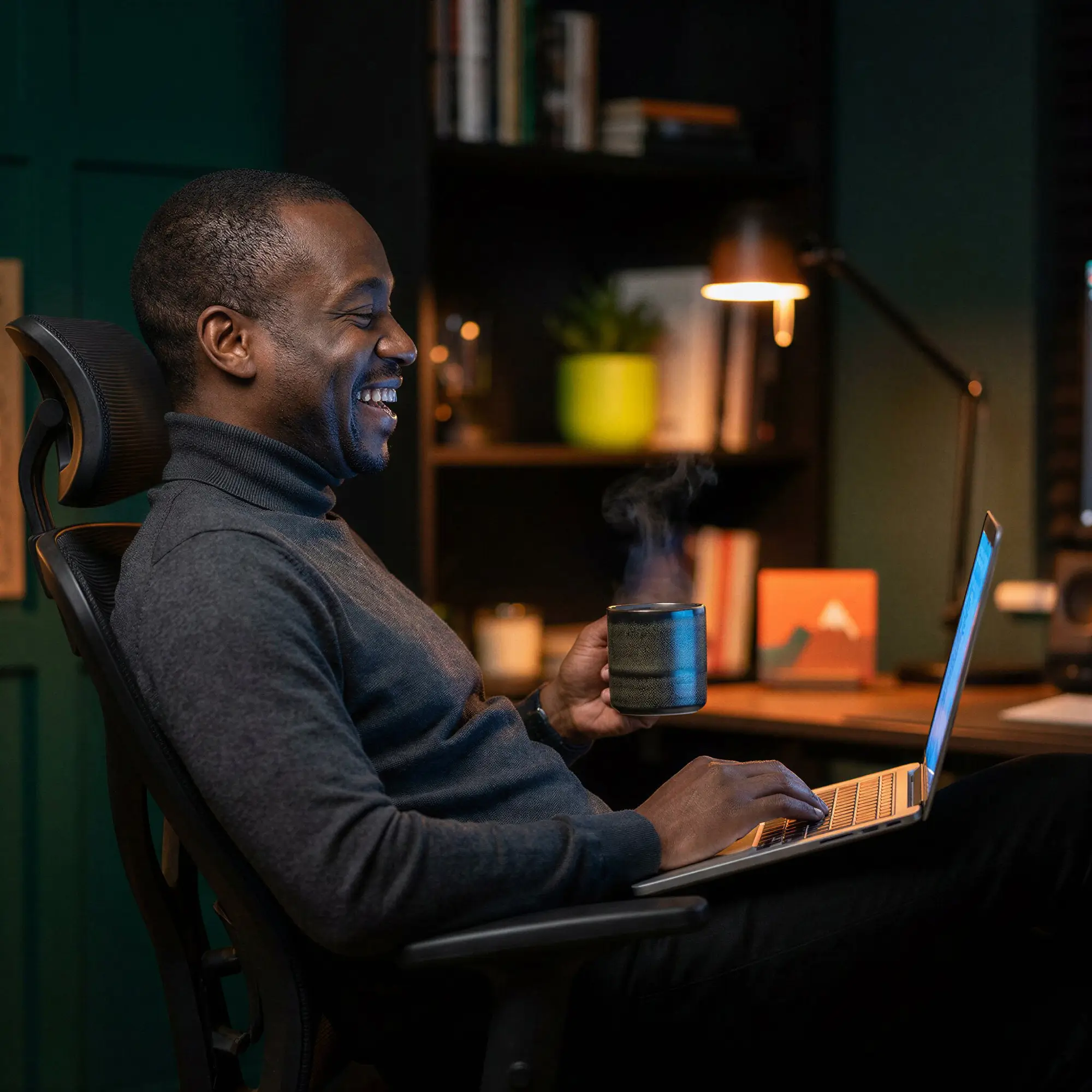 Smiling man sitting in an office chair holding a steaming mug and using a laptop in a cozy, dimly lit room.