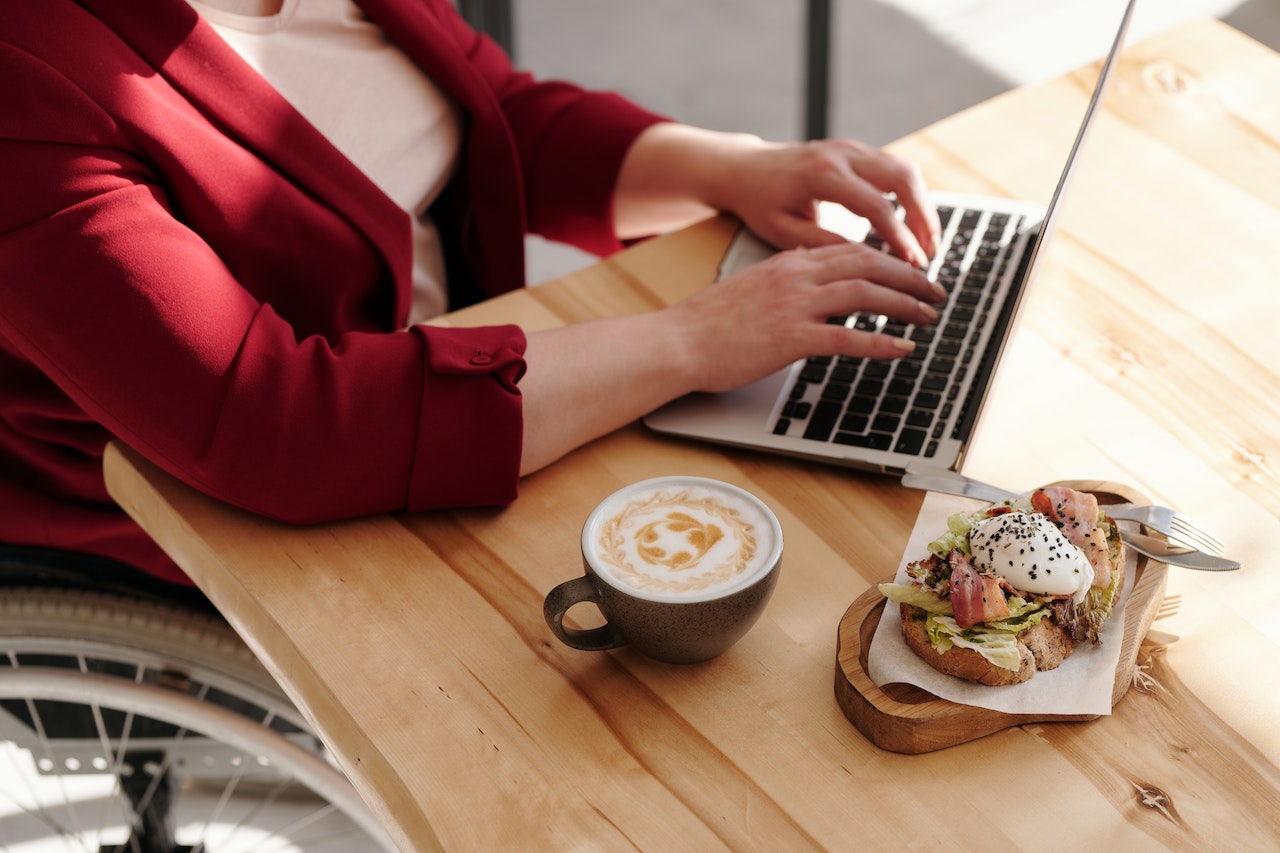 Close-up of a person in a red blazer typing on a laptop at a wooden café table, with a latte art cappuccino and an open-faced sandwich on a wooden board beside them. The scene depicts a remote virtual assistant working flexibly from a coffee shop environment.