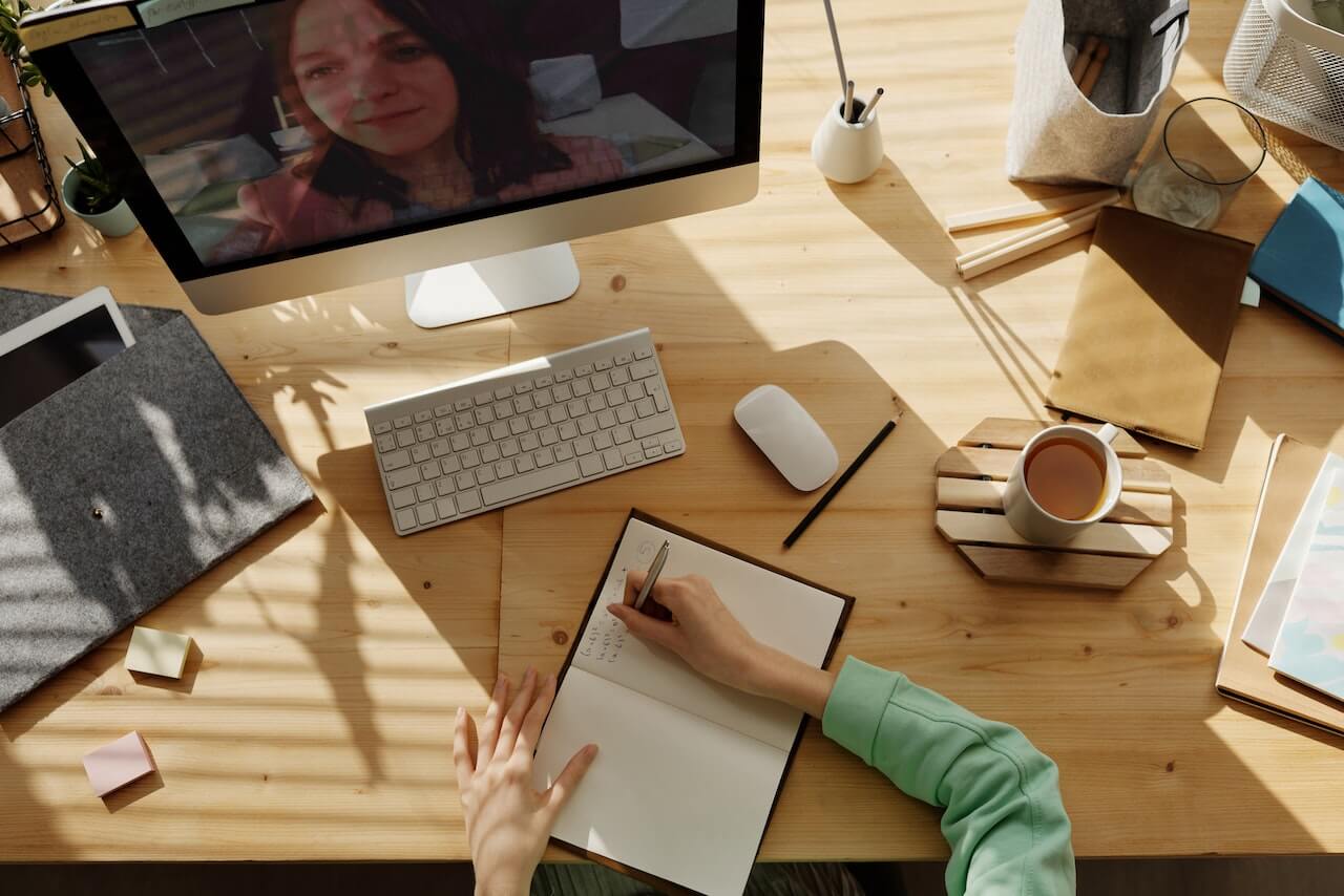 Overhead view of a sunlit wooden desk where a person in a green sweater takes notes in an open notebook during a video call on an Apple iMac. The screen shows a woman participant. The desk is organized with an Apple Magic Keyboard, Magic Mouse, sticky notes, a cup of coffee, pencils, and a tablet.