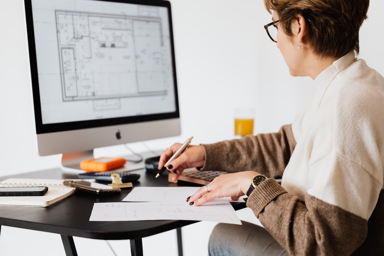A woman with glasses and short hair reviews architectural blueprints on paper while seated at a desk with an iMac displaying a floor plan, a calculator, and various office supplies. The image represents remote design or administrative roles in the architecture and real estate sector.