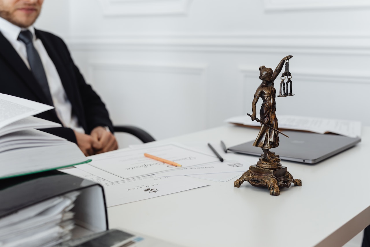 A man in a business suit sits behind a desk with legal documents and binders, with a Lady Justice bronze figurine in the foreground. The image represents the legal sector and the possibility of hiring remote legal administrative support through a Philippine staffing agency.
