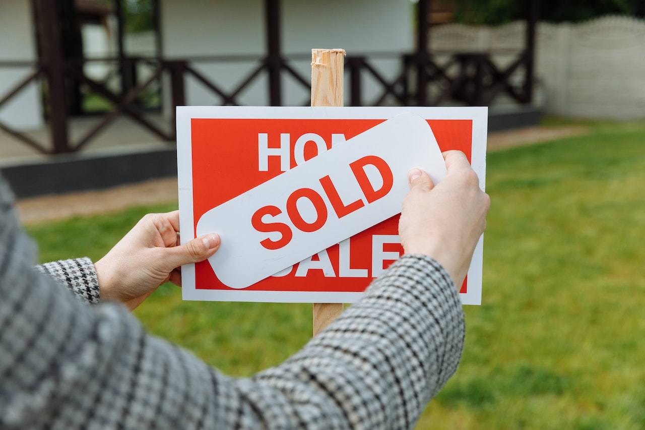 Hands placing a red "SOLD" sticker over a "Home for Sale" real estate sign in front of a residential property with a lawn. The image represents real estate transaction roles that can be filled by remote specialists hired through a Philippine headhunter agency.