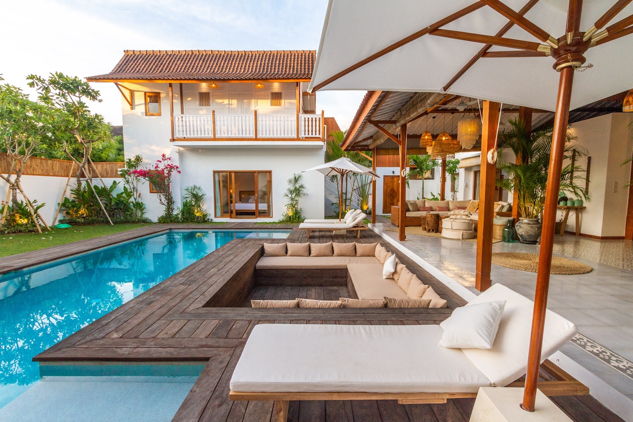 A luxurious tropical villa with a private swimming pool, wooden deck with cushioned sunbeds, outdoor lounge area, and a two-story white stucco house surrounded by lush landscaping. The image represents high-end real estate properties that remote real estate assistants can help manage and market.