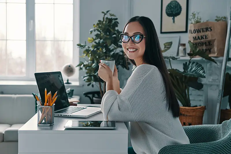 A smiling young woman with long dark hair and cat-eye glasses turns toward the camera while holding a coffee cup at her home office desk. Her laptop and pencil holder are visible on the white desk, with lush indoor plants and decorative wall art in the bright background, depicting a relaxed and productive remote work environment.