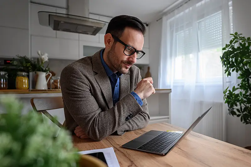 A focused man wearing glasses and a blazer leans forward at a wooden kitchen table, studying content on his laptop with a thoughtful expression and his fist raised to his chin. Plants, a white kitchen, and natural light from a nearby window create a calm remote work setting, illustrating a business professional evaluating nearshore outsourcing options.