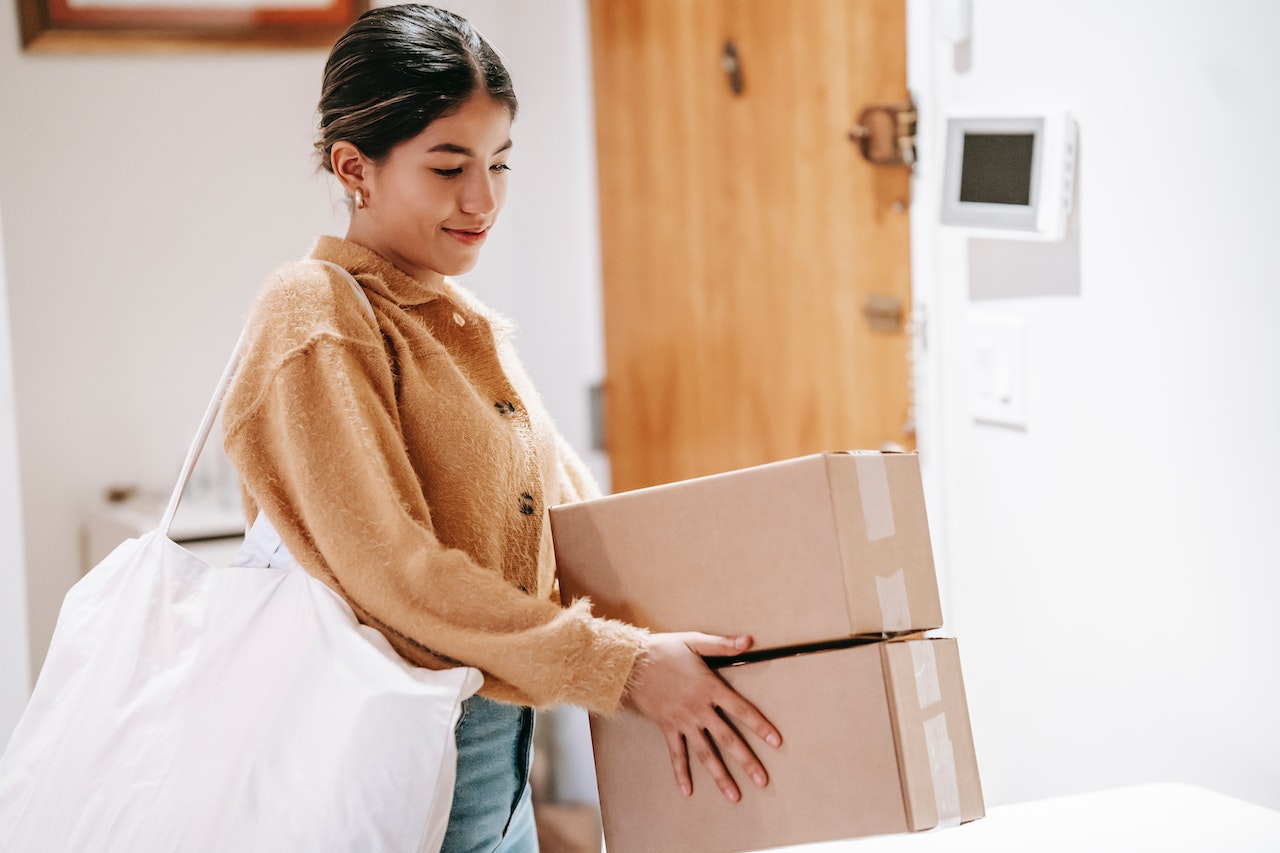 A young woman in a mustard-yellow sweater carries two stacked cardboard boxes while also holding a tote bag, standing in a bright indoor space near a thermostat on the wall. The image represents operations, logistics coordination, or an office manager role.