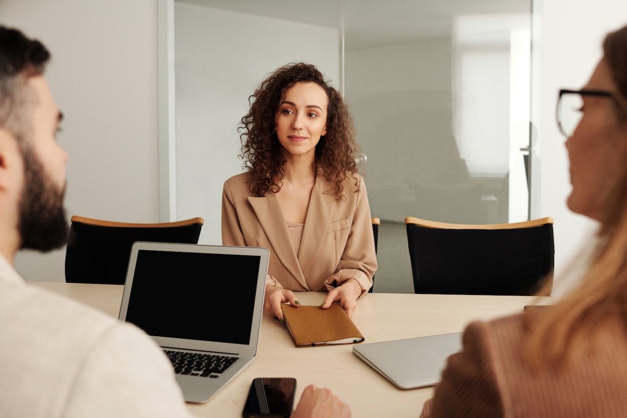 A woman with curly hair in a beige blazer sits confidently across a conference table from two interviewers, holding a notebook. She represents a leadership candidate being evaluated for a remote executive or managerial position.