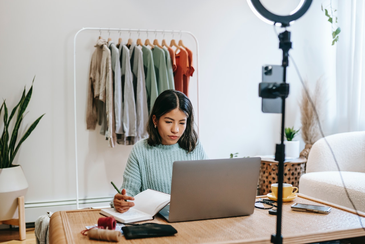A young woman with dark hair sits at a home desk with a laptop and ring light setup, taking notes in a notebook against a backdrop of hanging clothing on a rack. She represents a remote content manager, e-commerce manager, or social media director working from home.