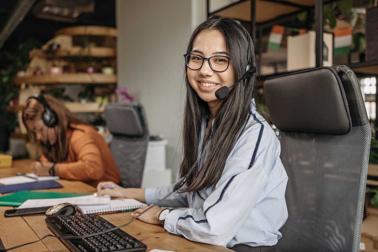 A young Asian woman wearing glasses and a headset smiles at the camera while seated at a desk with a keyboard and mouse in a plant-decorated office. A colleague in an orange top is seen working in the background, representing a customer support lead or operations role.
