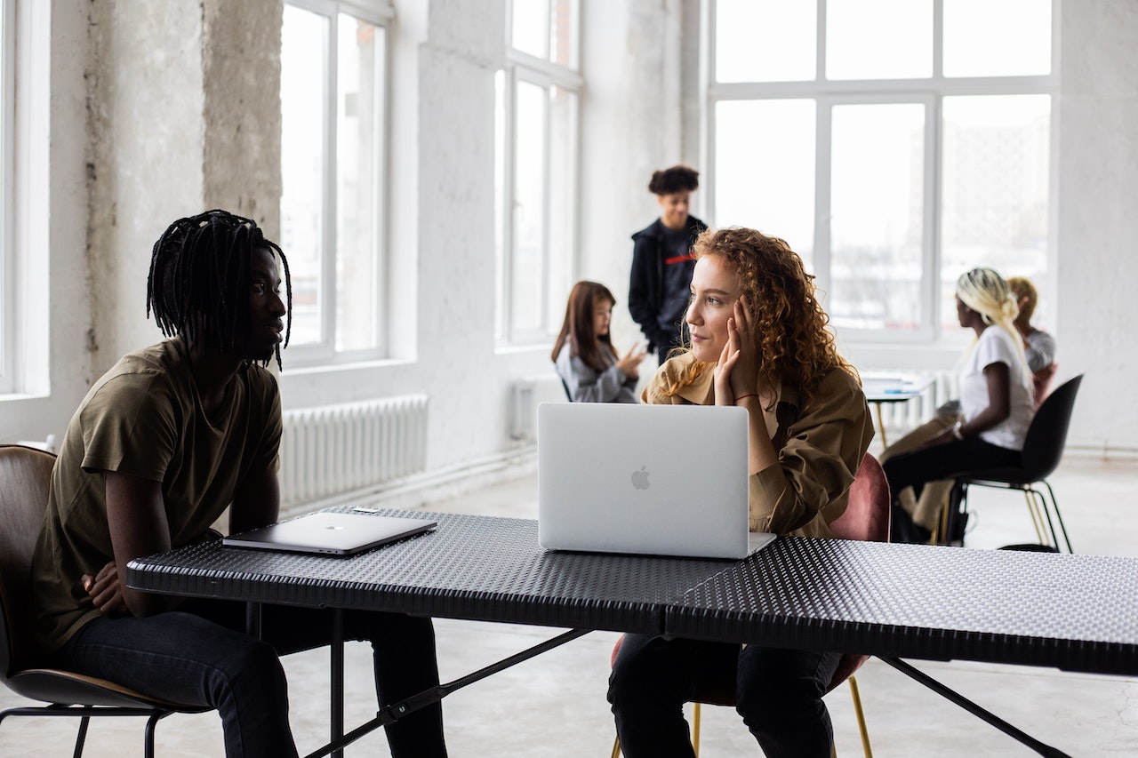 A diverse group of remote workers sits at a long table in a bright, industrial-style open workspace. Two people in the foreground — a man with dreadlocks and a woman with curly red hair — appear to be having a direct conversation, with other colleagues visible working in the background.
