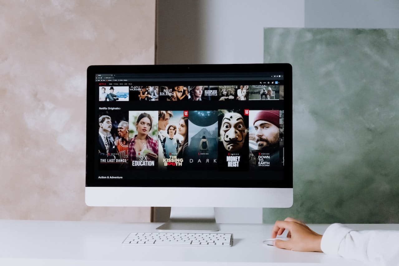 A desktop computer monitor on a white desk displays a Netflix browsing interface showing titles including The Last Dance, Sex Education, The Kissing Booth, Dark, Money Heist, and Down to Earth. A hand holds a mouse beside a keyboard, illustrating a virtual team watch party activity.
