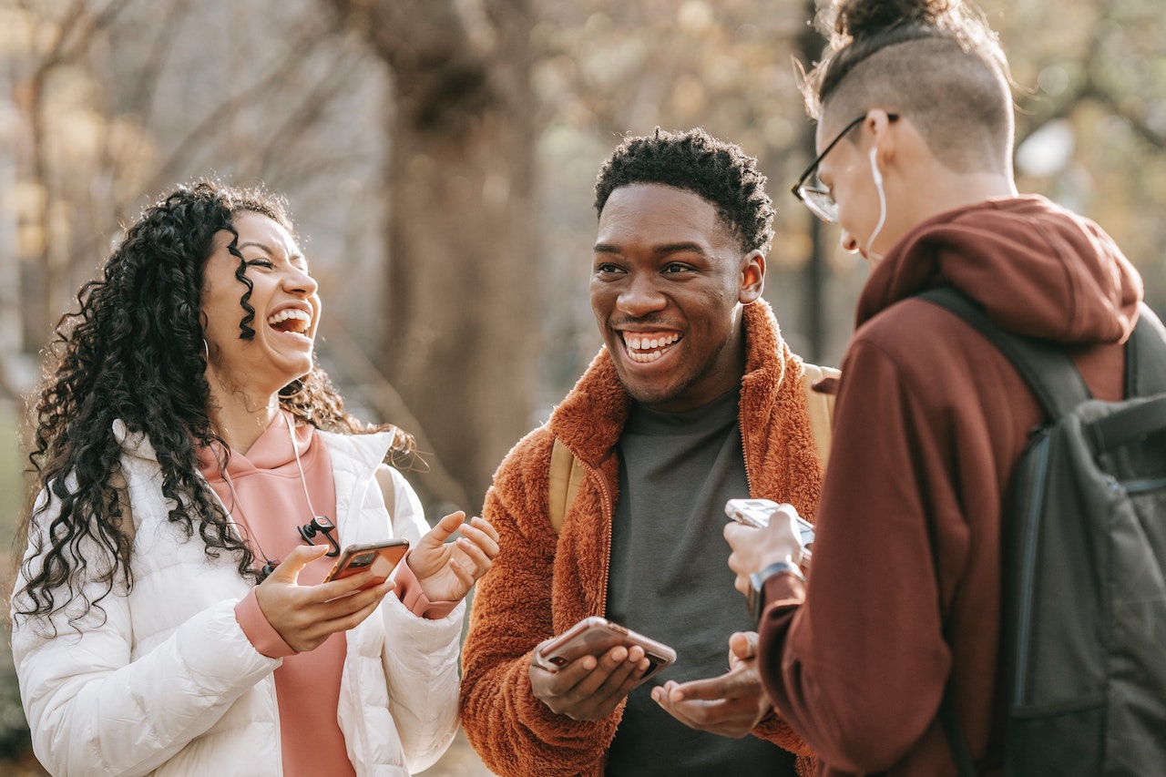 Three young people laughing and chatting outdoors in a park setting, each holding a smartphone. Two face forward while one stands with their back to the camera. The scene conveys friendly, open communication — illustrating the interpersonal and social soft skills valuable in a virtual assistant.