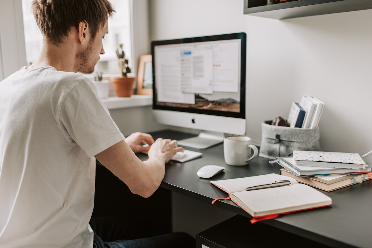A young man in a white t-shirt seen from behind, sitting at a home office desk and typing on an Apple iMac. The desk holds an open notebook, a coffee mug, a mouse, and stacked books. A window with plants sits behind the monitor, depicting a focused remote work environment.