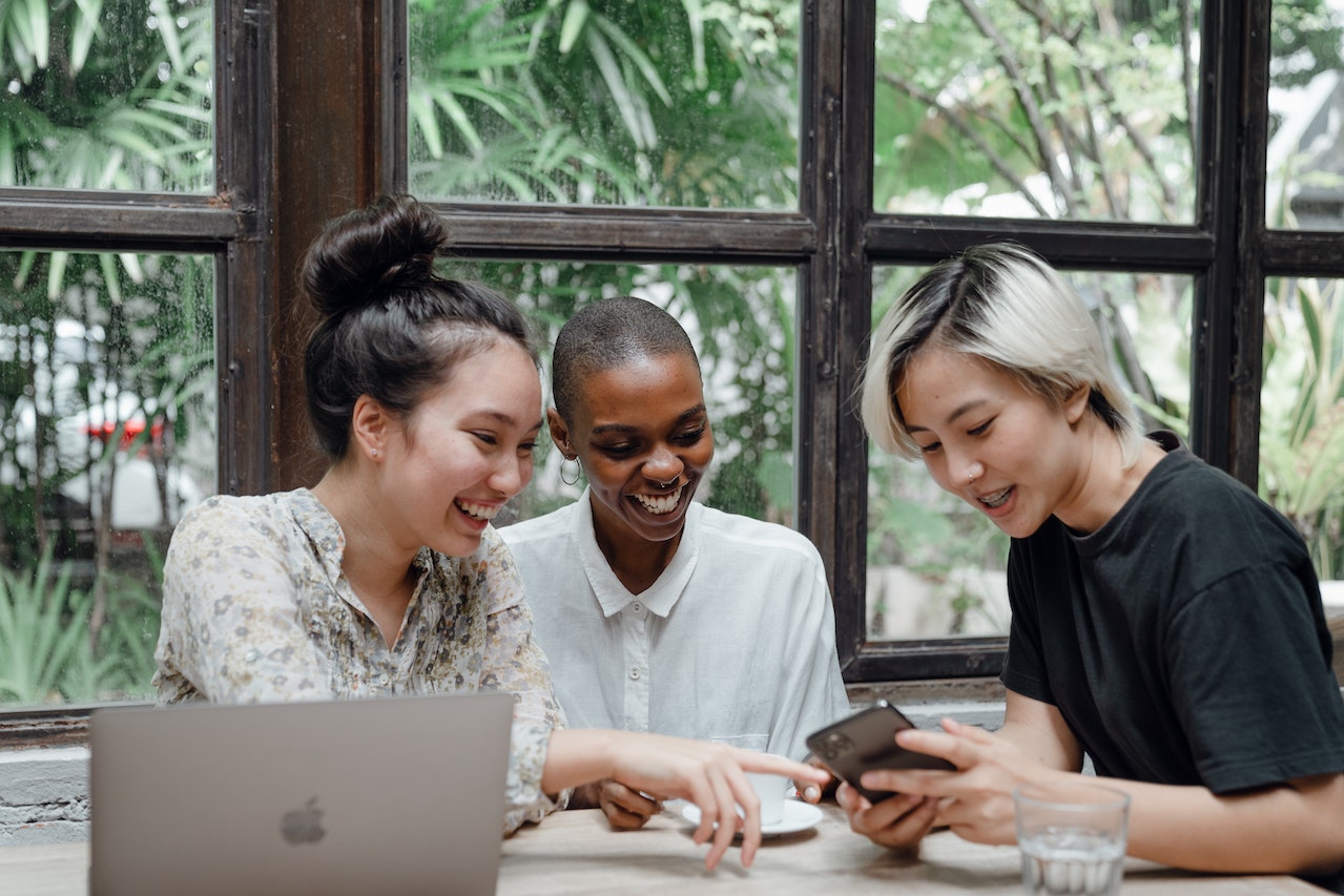 Three diverse young women sitting together at a cafe table, smiling and looking at a smartphone screen being held by one of them. A laptop is open in front of them and lush greenery is visible through large windows behind them, representing collaborative teamwork and communication skills.