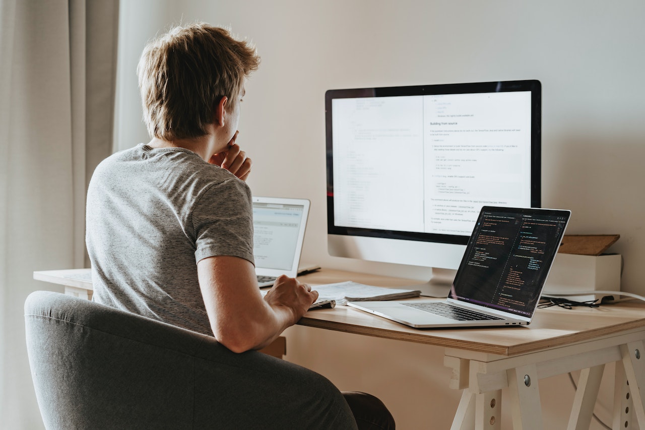 A remote worker viewed from behind, seated at a wooden desk with a large external monitor displaying a document and a laptop showing code on a dark background. The setup suggests a developer or technical professional working from a home office.