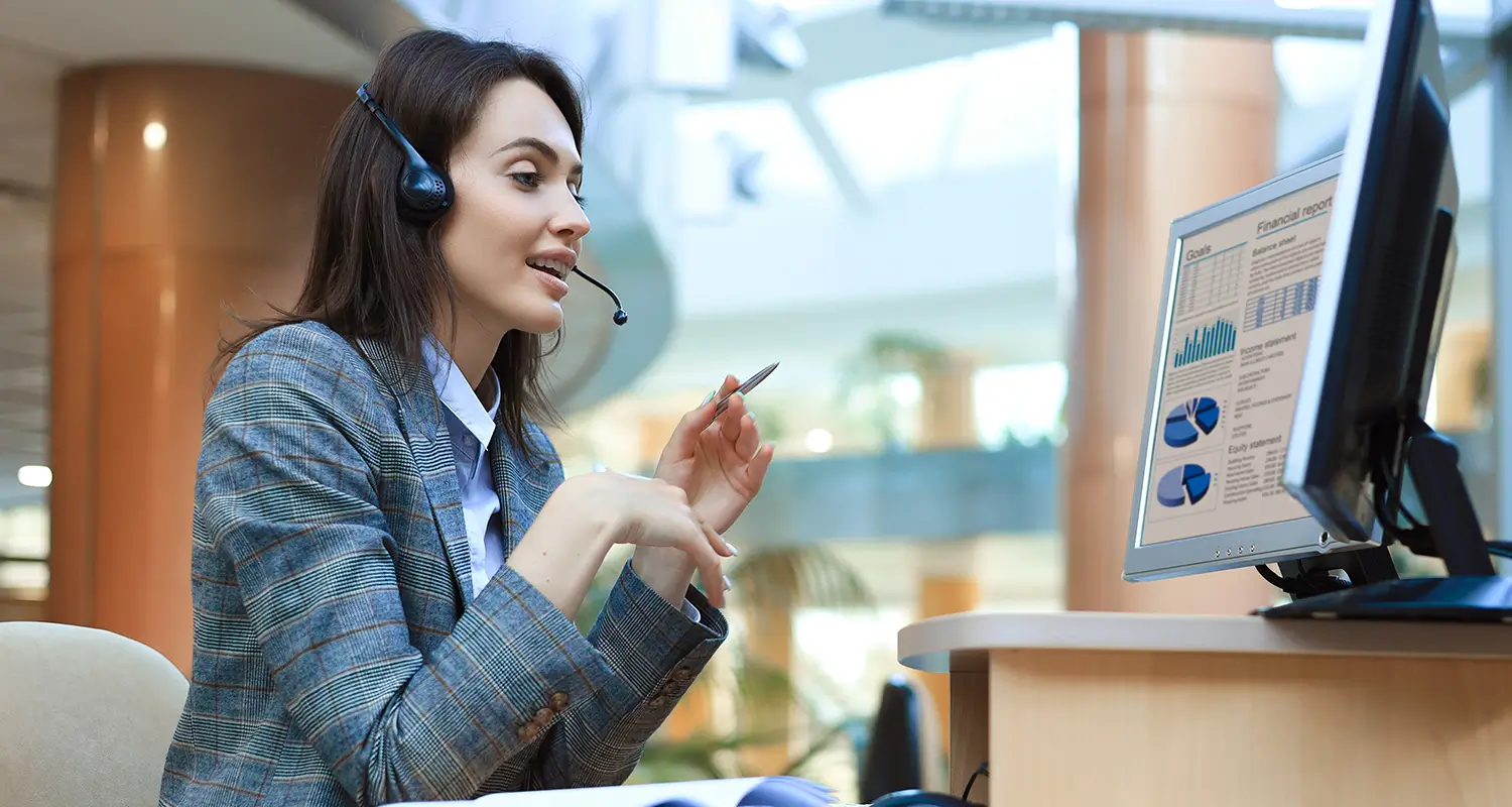 A professional woman wearing a headset and checkered blazer gestures with a pen while speaking at her computer in a bright modern office. Her monitor displays a financial report with pie charts and bar graphs, illustrating a BPO sales representative managing client accounts and driving revenue outcomes remotely.