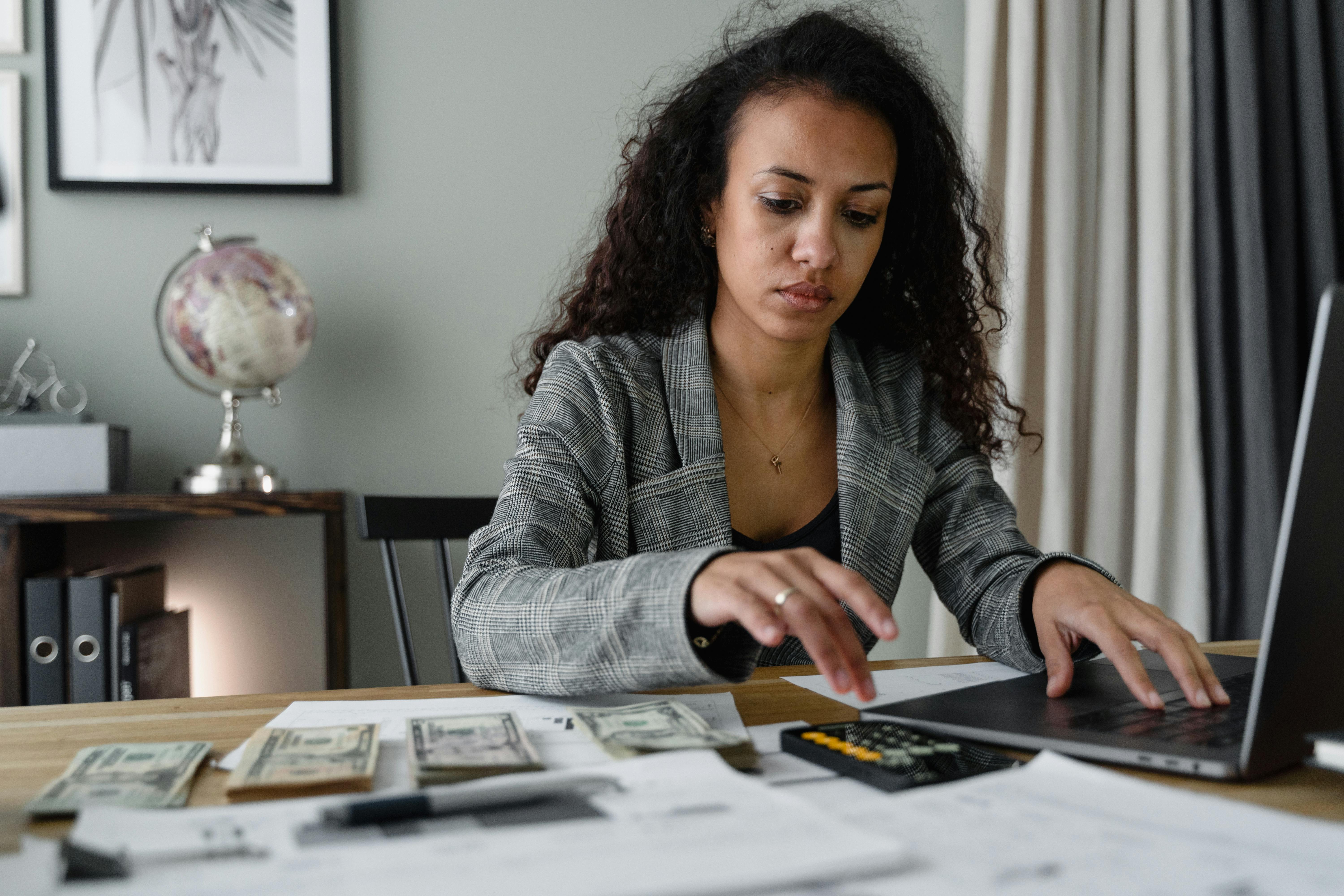 Woman in blazer working on laptop with calculator and cash on desk, managing finances