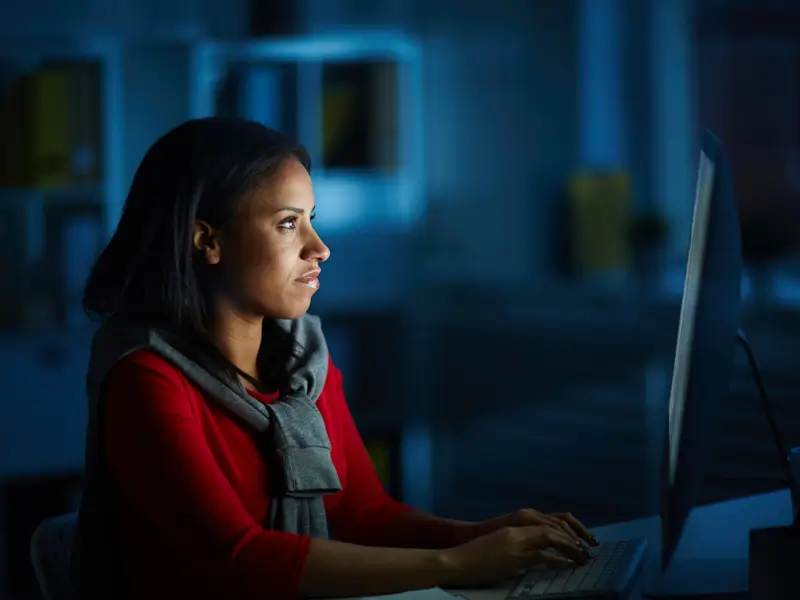 A woman in a red top and grey scarf sits at a desktop computer in a darkened office, her face illuminated by the blue glow of the monitor screen. Her focused expression and late-night setting suggest a developer or technical professional working concentrated hours on a demanding project.