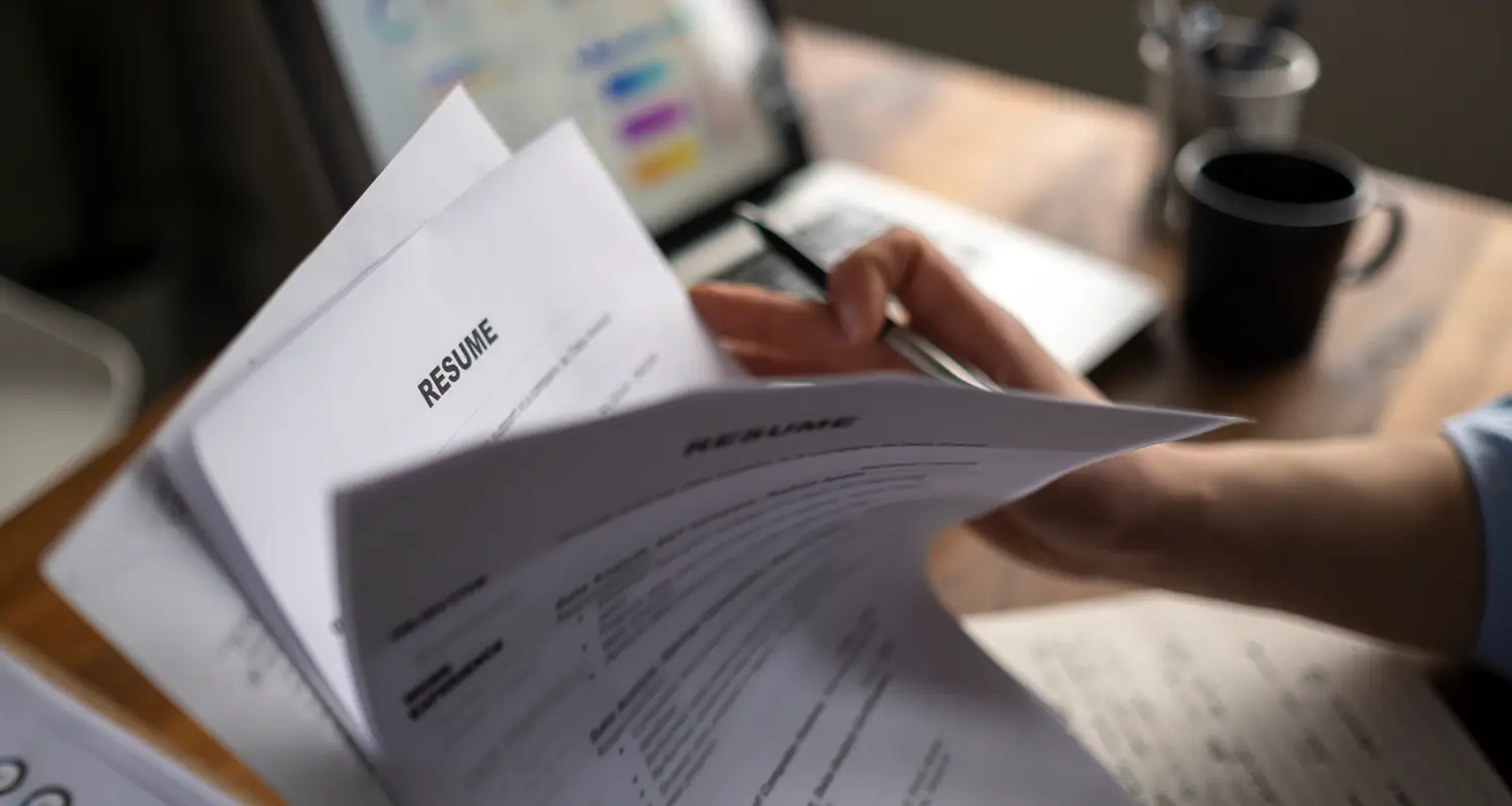 A person holds a stack of multiple resumes labeled "RESUME" while sitting at a desk with a laptop and coffee mug visible in the blurred background. The image illustrates the overwhelming volume of applications recruiters face and reinforces the argument for skills-first hiring practices that look beyond impressive resumes.