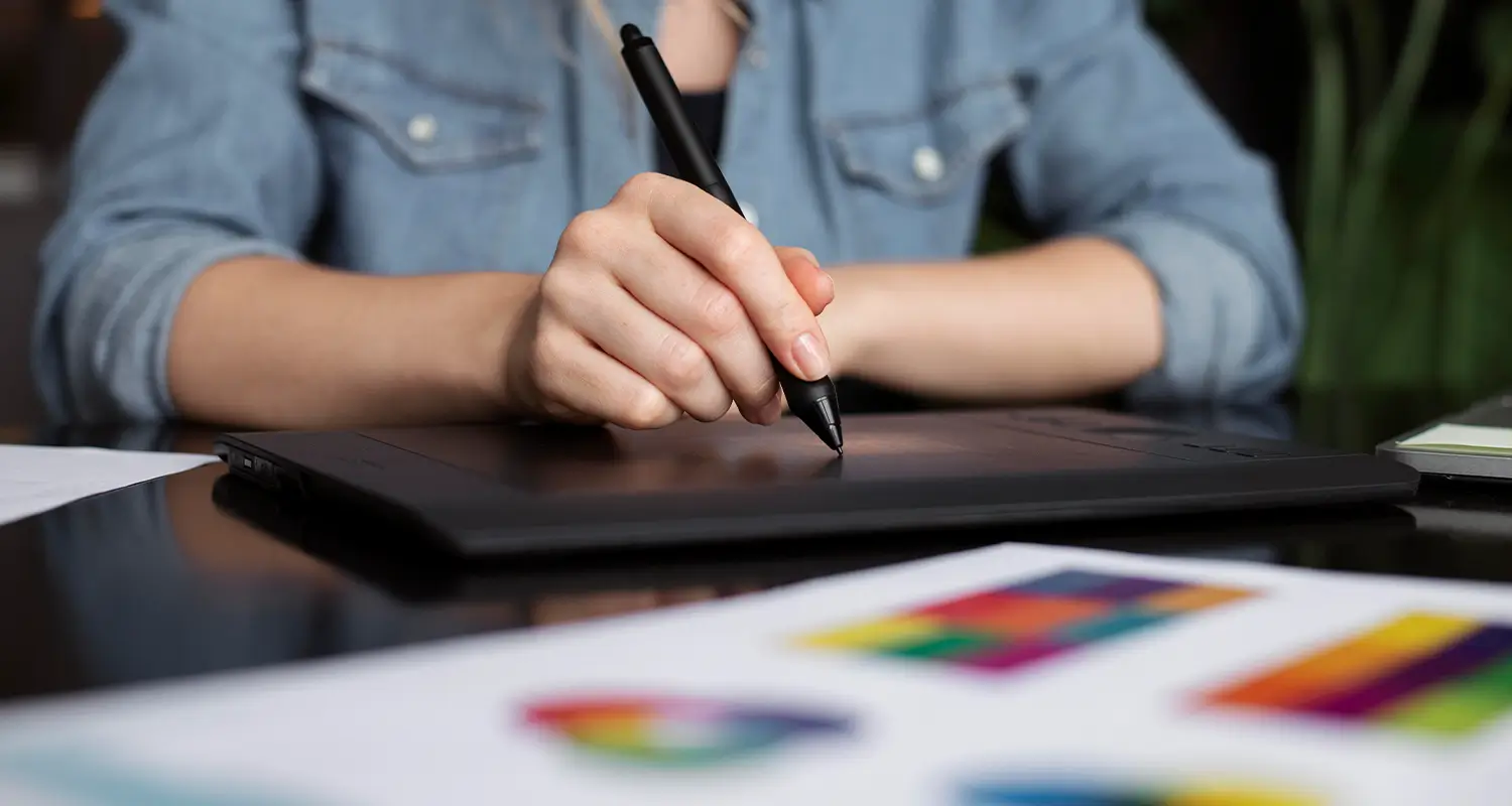 A close-up of a person's hand using a stylus pen on a graphics tablet, with color swatch cards fanned out in the foreground. The image illustrates demonstrated creative skill in action, representing the skills-first hiring philosophy where practical ability matters more than credentials listed on a resume.