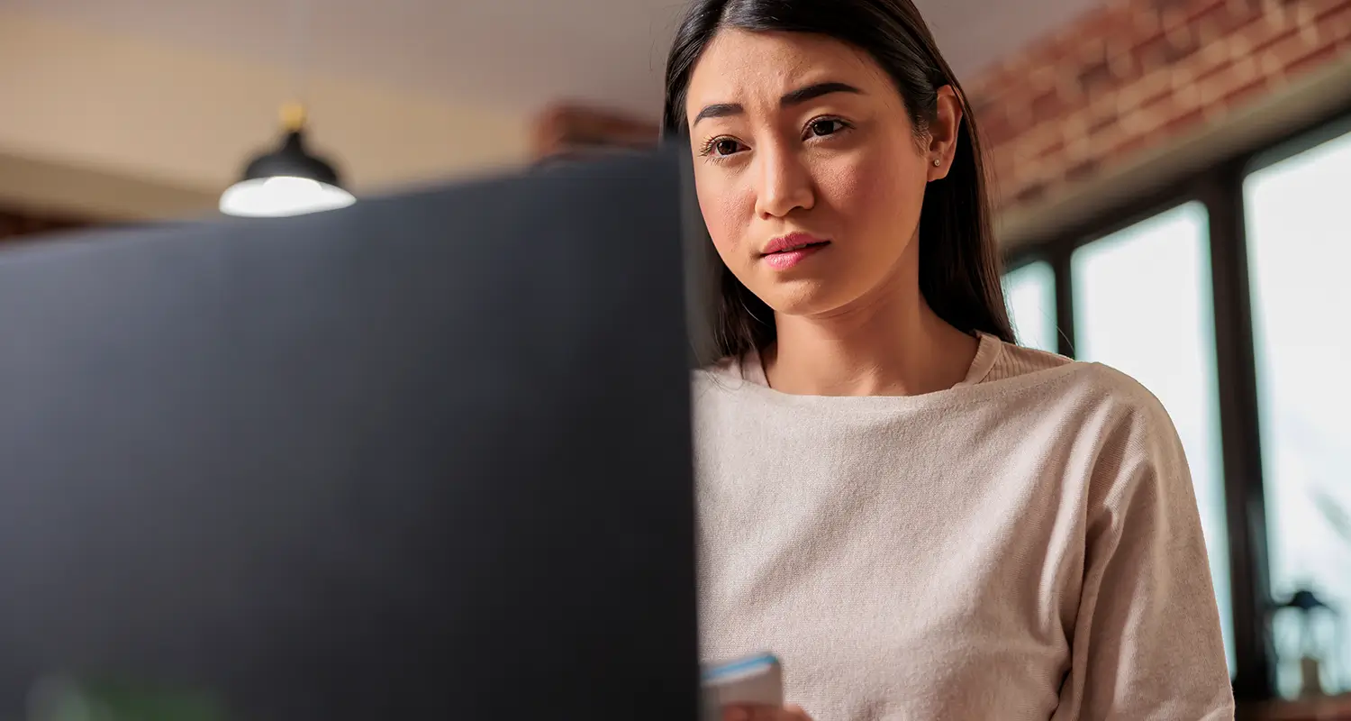 Concerned Asian woman with straight hair staring at a large monitor screen with a worried expression, holding a phone in a dim home office.
