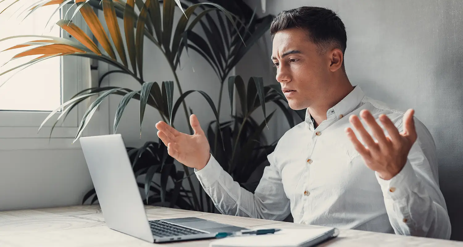 Frustrated young man in a white shirt gesturing with open hands at his laptop screen, looking confused and disengaged while working remotely.
