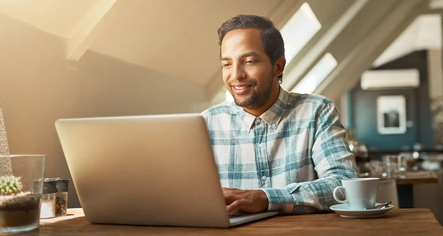 A smiling man in a blue plaid shirt works on a laptop at a wooden cafe table bathed in warm sunlight, with a coffee cup and small cactus plant beside him, conveying a relaxed and productive remote work atmosphere.