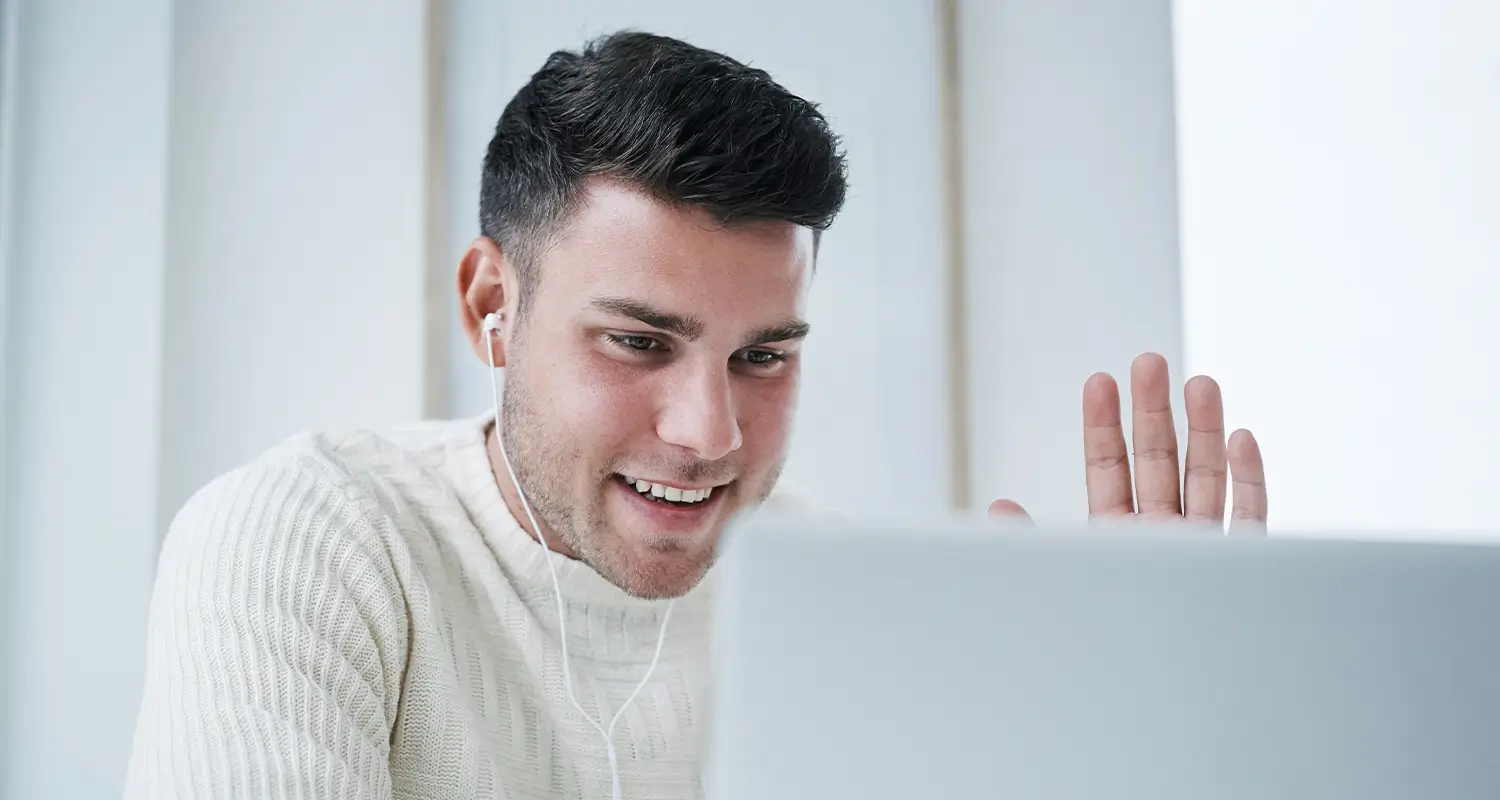 A young man in a cream sweater wearing white earbuds waves at his open laptop screen with a smile, engaged in a video call, set against a bright, minimalist background suggesting a home office environment.