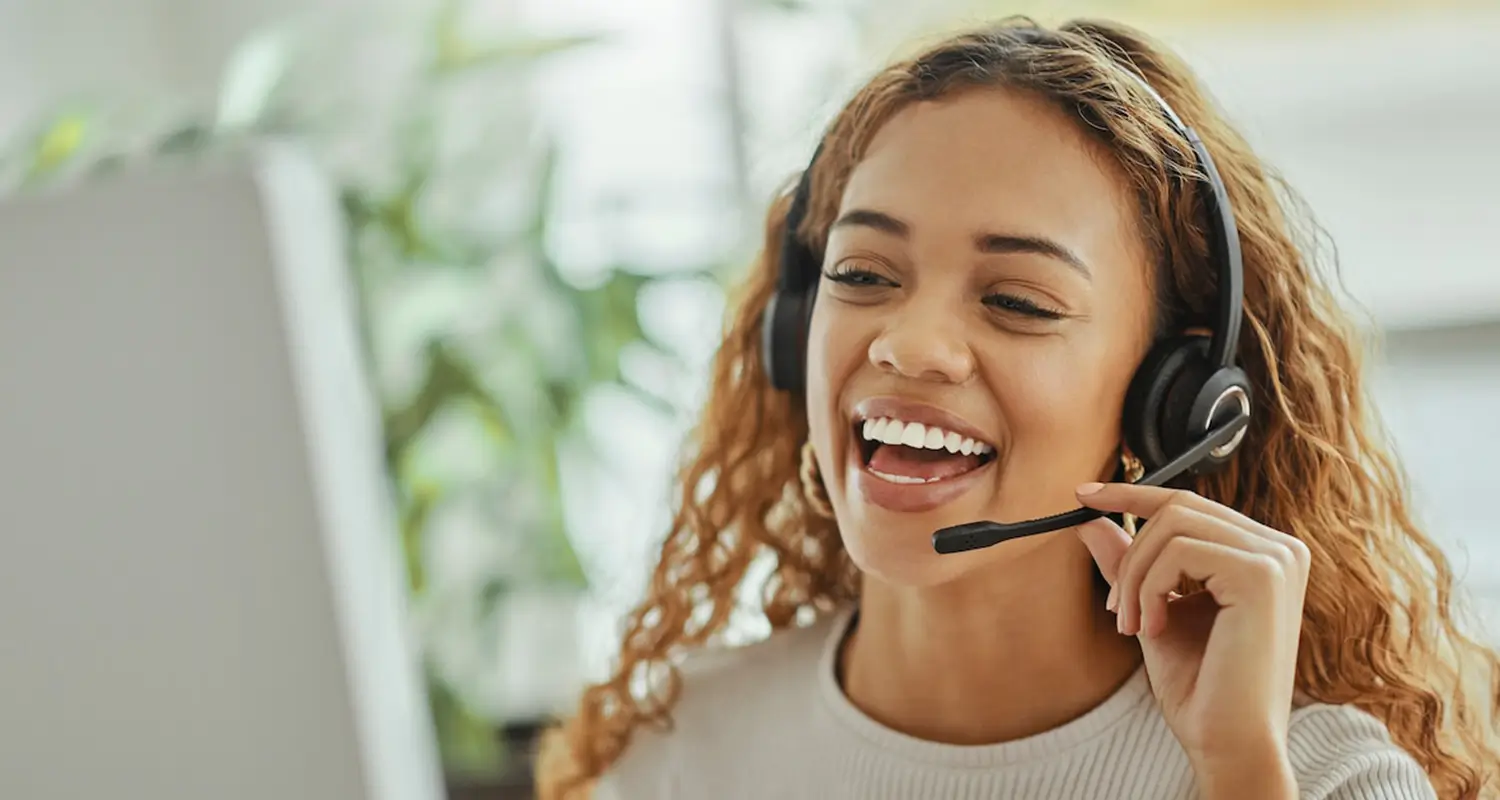 A young woman with curly auburn hair and a headset laughs while speaking with a customer, seated in a light-filled office with plants visible in the background. Her natural warmth and enthusiasm represent the kind of engaging offshore customer support representative who keeps satisfaction high and response times fast.