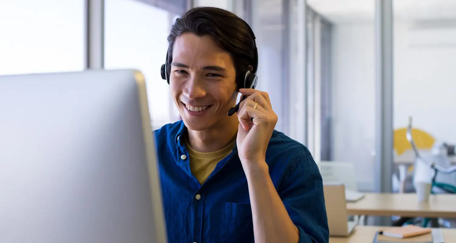 A smiling young Asian man wearing a headset holds the microphone while sitting in front of a desktop computer in a bright, modern office. His warm, attentive expression reflects the customer-focused communication skills that make offshore customer support representatives effective at resolving issues and building positive client relationships.