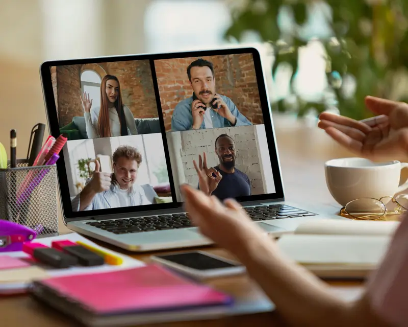 A laptop screen shows a video call with four remote team members—a woman waving, a man on a headset, a man giving a thumbs up, and a man making an OK sign—while a person at the desk gestures with their hands. The setup, surrounded by office supplies and a coffee mug, illustrates a team leader conducting a remote team meeting.