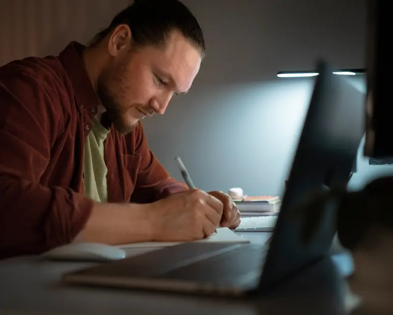 A bearded man in a dark red shirt writes notes in a notebook at a dimly lit desk, with an open laptop and a small desk lamp illuminating his workspace. The focused, close-up scene conveys a leader planning strategy or documenting team goals during a late work session.