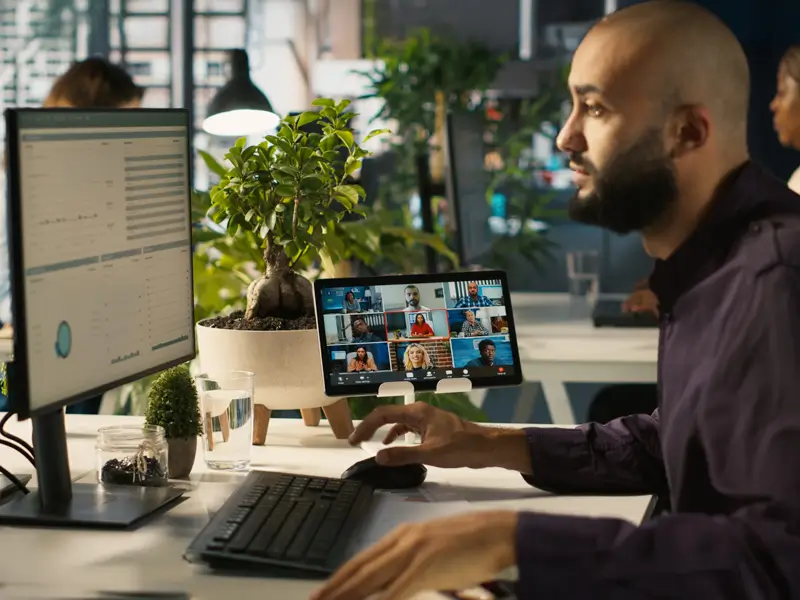 Bearded man at a dual-monitor desk joining a video call with a diverse group of remote team members on a tablet