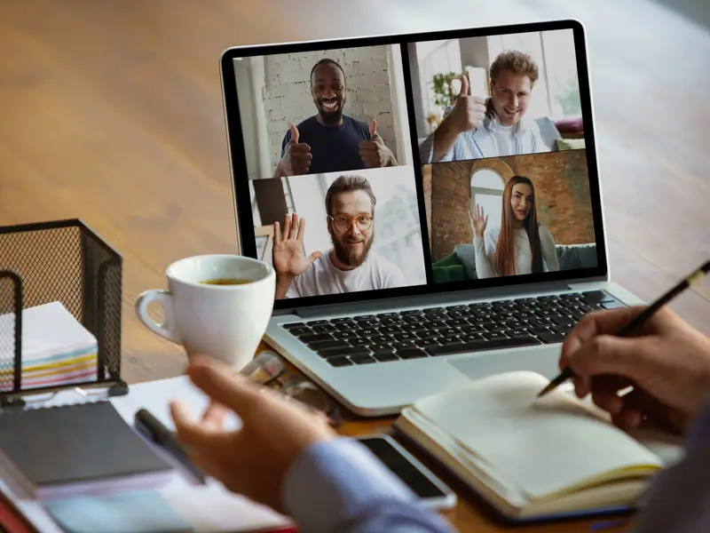 Person taking notes in a journal while on a laptop video call with four smiling remote team members waving