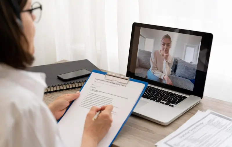 Viewed from behind, a woman with dark hair and glasses writes notes on a clipboard while conducting a video interview on a laptop. The laptop screen shows a young woman sitting casually on a sofa, representing a remote hiring or follow-up interview scenario where the interviewer takes structured notes during the session.