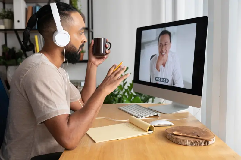 A bearded man wearing white over-ear headphones sits at a wooden home-office desk, holding a dark coffee mug in one hand and a pencil in the other, as he attends a video call. The desktop monitor shows a smiling man on the call, suggesting a remote follow-up interview or one-on-one hiring conversation in a casual home setting.