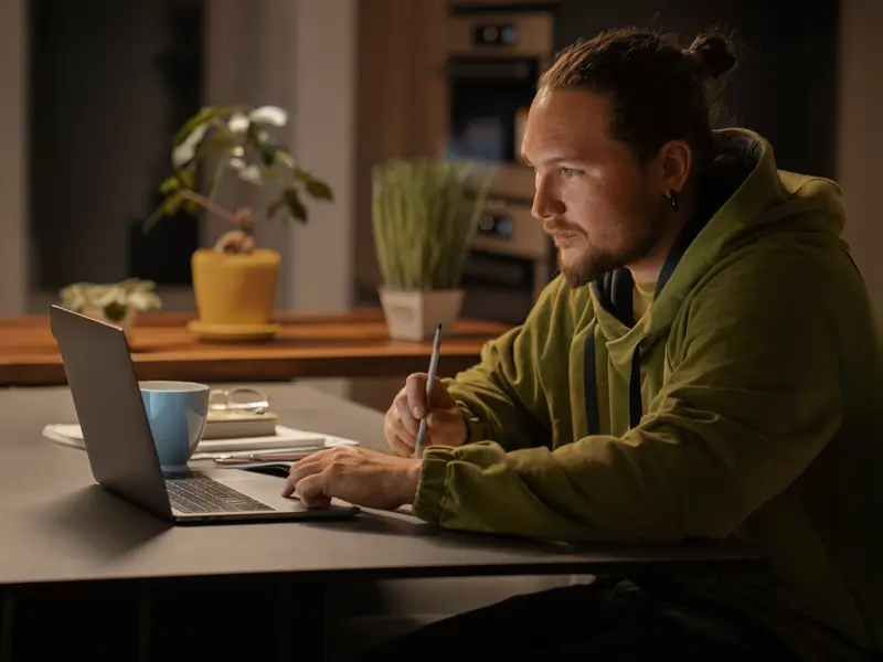 A young man with a bun hairstyle and headphones around his neck sits at a kitchen table in dim evening lighting, working on a laptop and taking notes with a pen. A blue mug and plants are visible in the background, suggesting a focused remote work or research session.