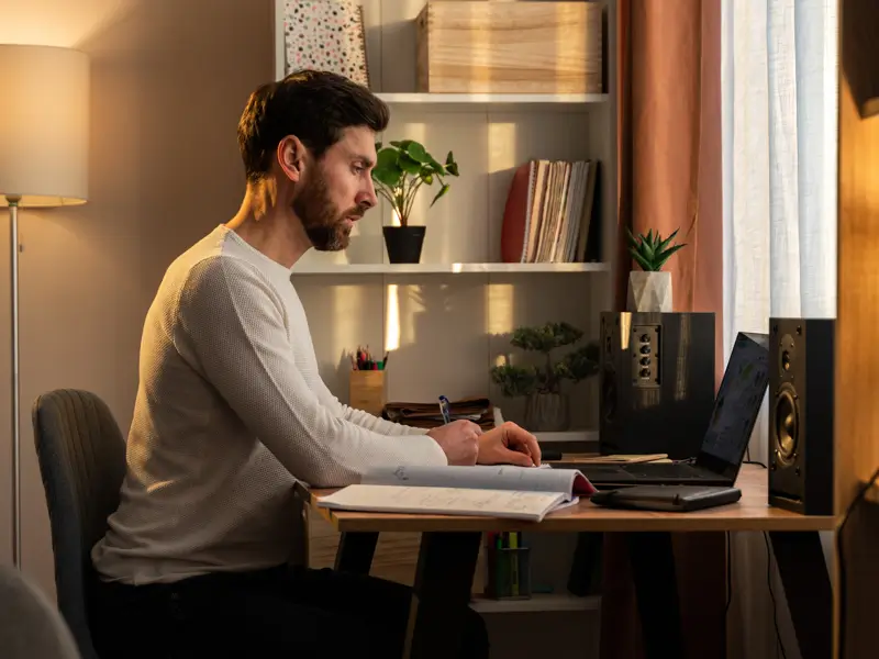 A bearded man in a white sweater sits at a home office desk, focused on an open notebook with a laptop beside him. A bookshelf filled with records and plants is visible behind him, along with a floor lamp casting warm light, conveying a productive remote work environment.