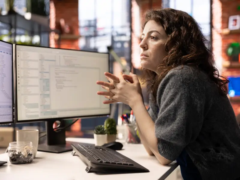 A woman with curly dark hair sits at a desk in a modern office space, gazing thoughtfully at two large monitors displaying code or data. Her hands are clasped in front of her as she reviews the screens, with brick walls and plants visible in the background.