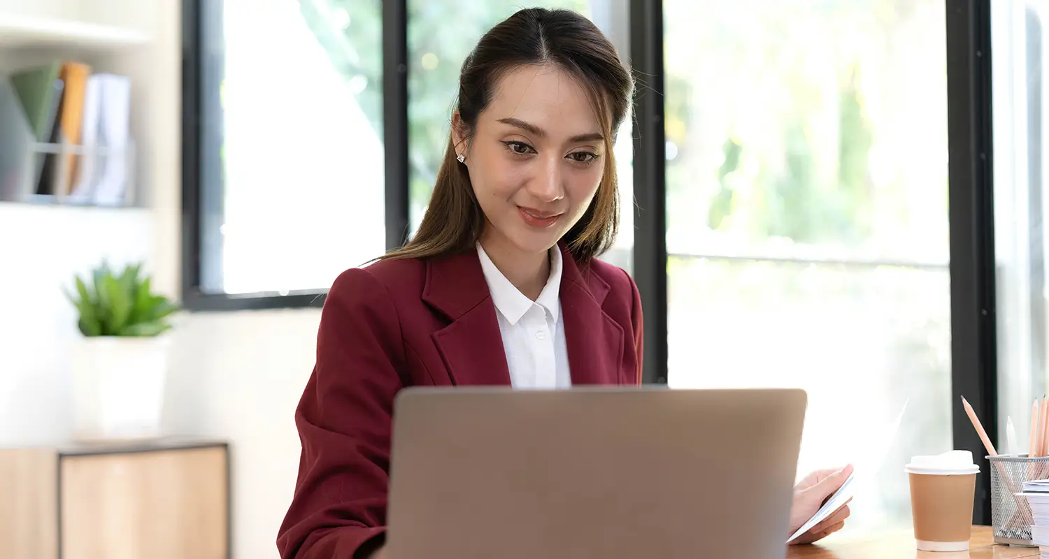 A young Asian woman in a red blazer smiles while working on a laptop in a bright, plant-filled office. A coffee cup and pencil holder sit nearby on the clean desk, representing an offshore professional hired directly by a company and seamlessly integrated into their global team.