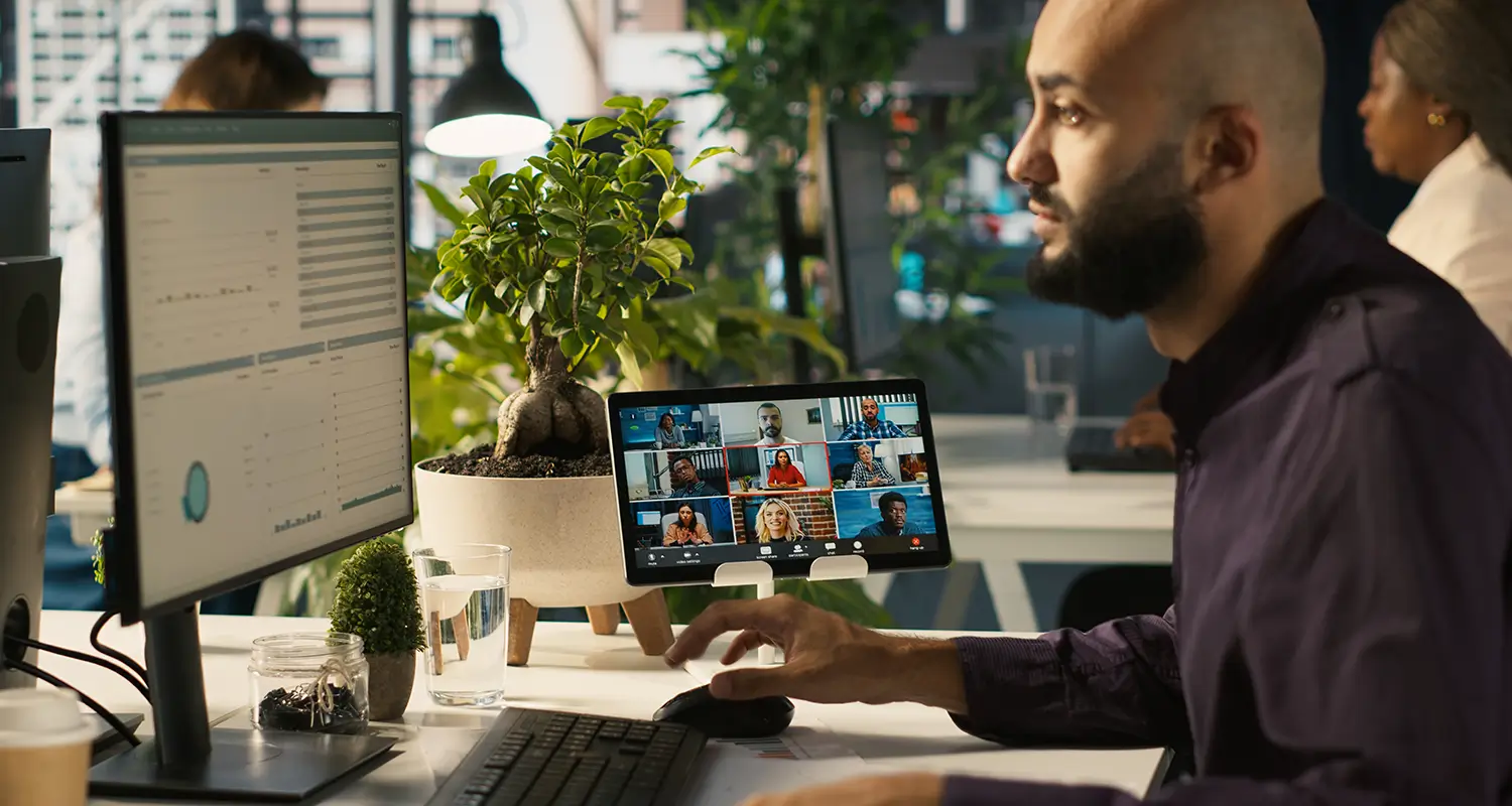 A bearded man in a dark purple shirt sits at a busy office desk with two monitors — one showing a data dashboard and another displaying a video conference grid with nine remote colleagues. The scene depicts a team manager equipped with the tools needed to coordinate and communicate across a globally distributed workforce.