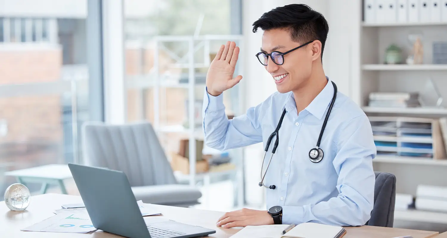 Doctor with stethoscope waving during a video call on a laptop in a bright office